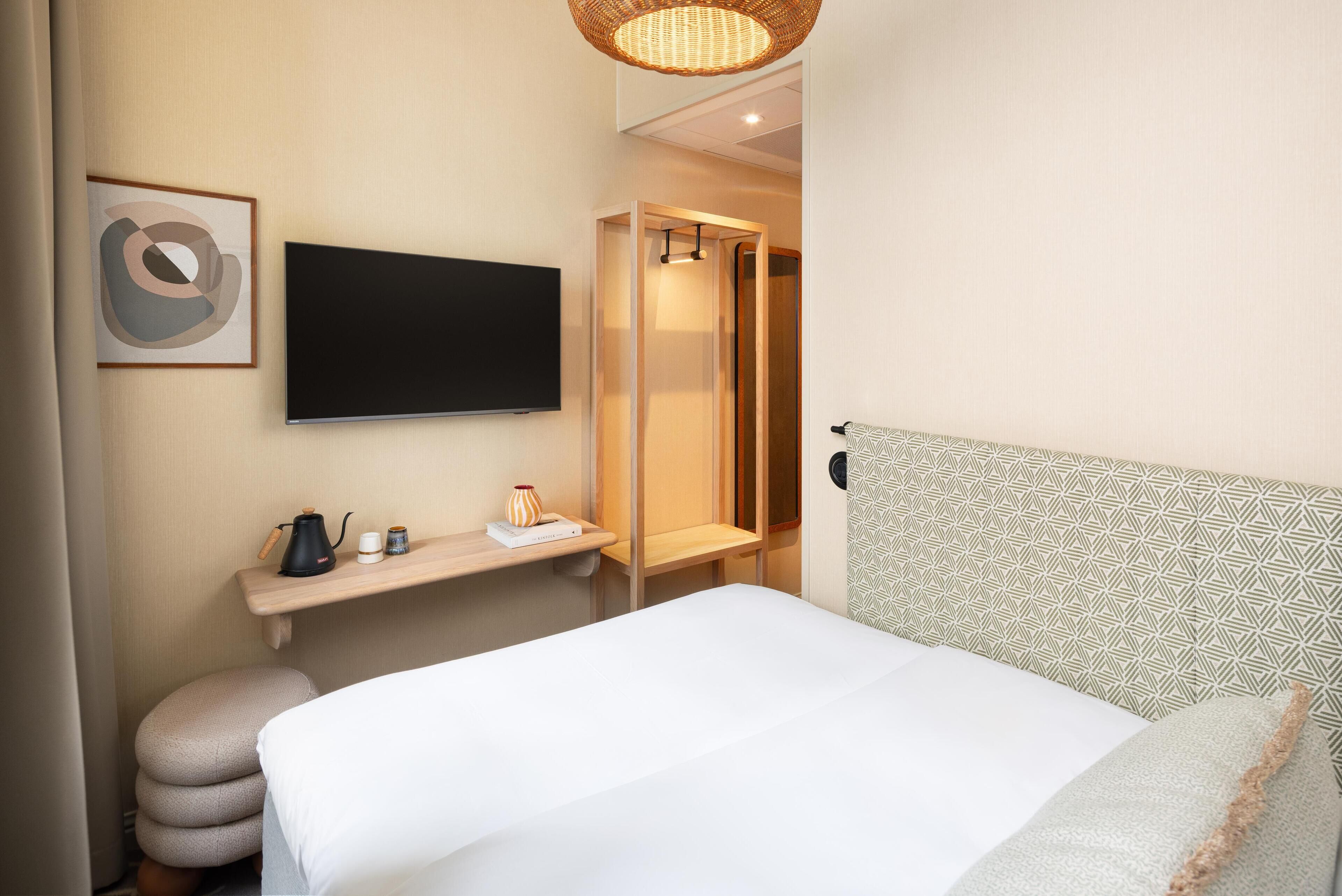 Habitación de hotel con cama, televisor montado en la pared, una pequeña estantería con hervidor y un jarrón, y un cabecero estampado.
