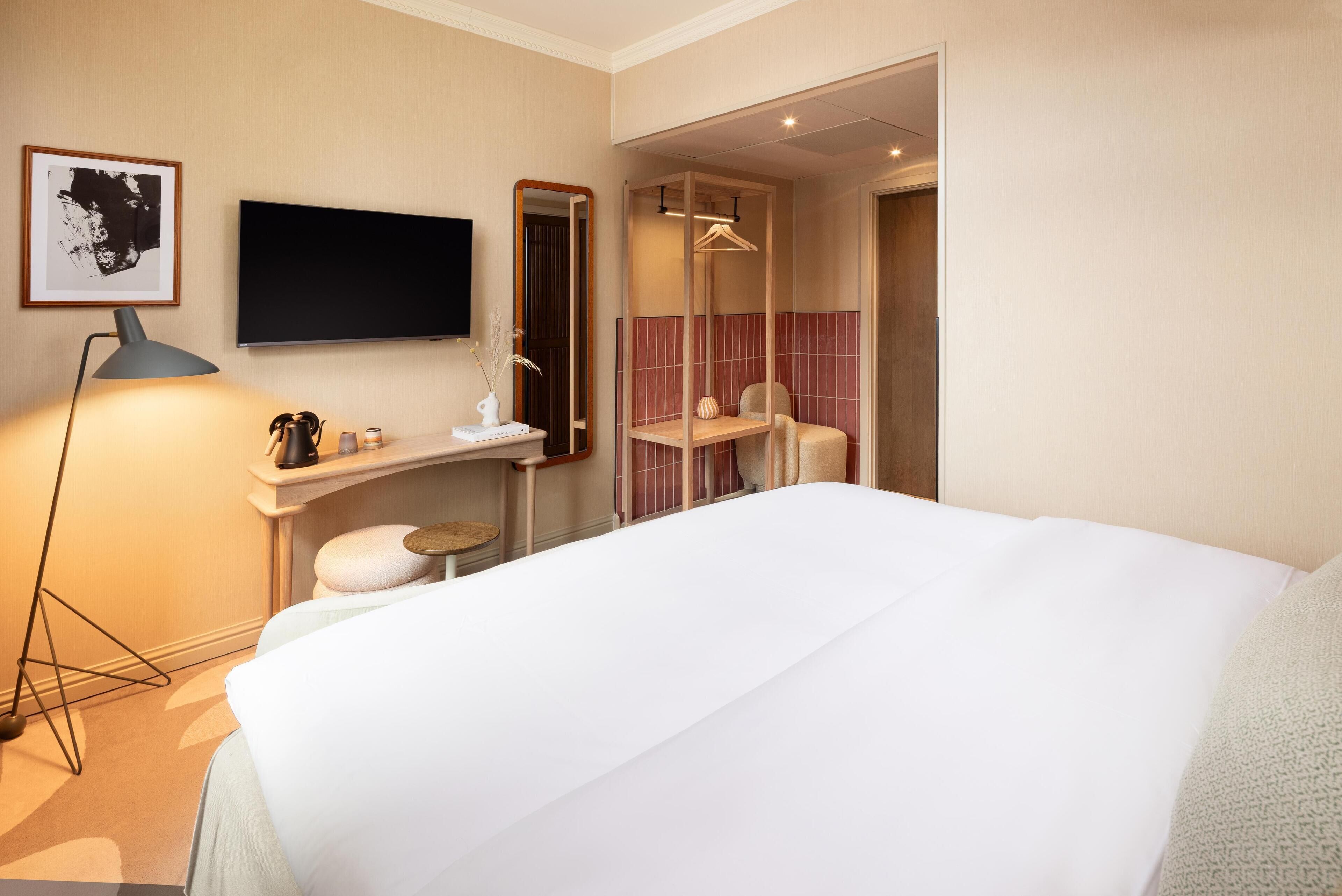 A hotel room with a bed, a desk, a mirror, a television, and a bathroom visible through an open door.
