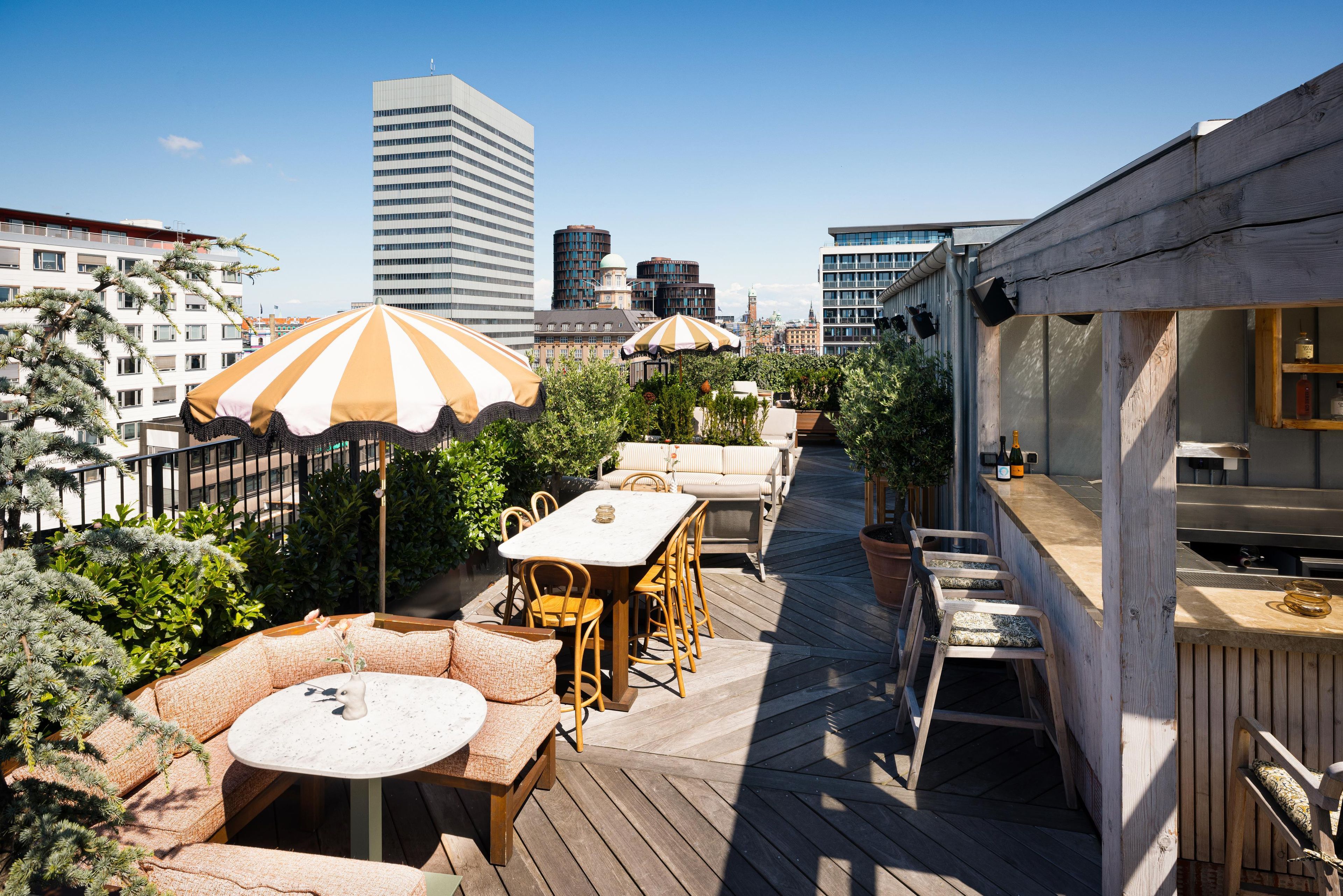 An outdoor deck with a dining area, including a table and chairs, surrounded by greenery and umbrellas.