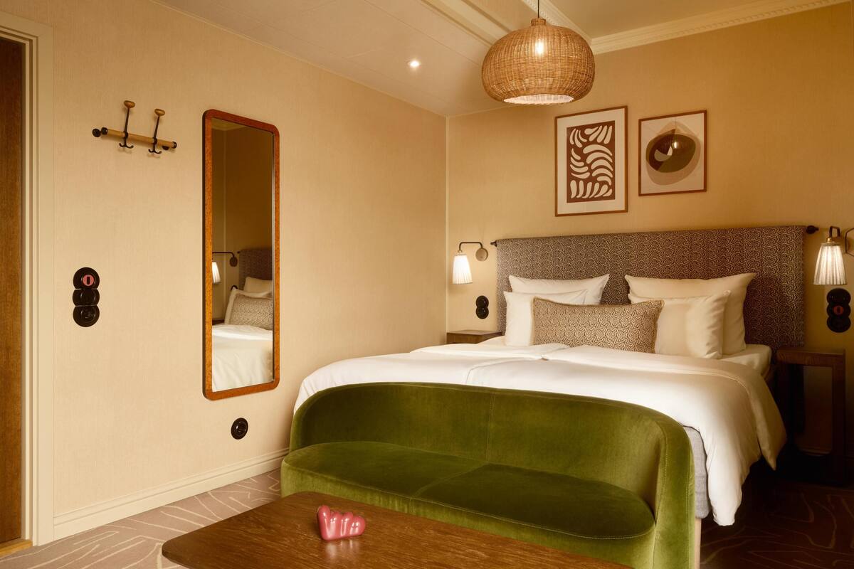 A hotel room with a large bed, a green sofa, a wooden table, a mirror, wall-mounted lights, and artwork.