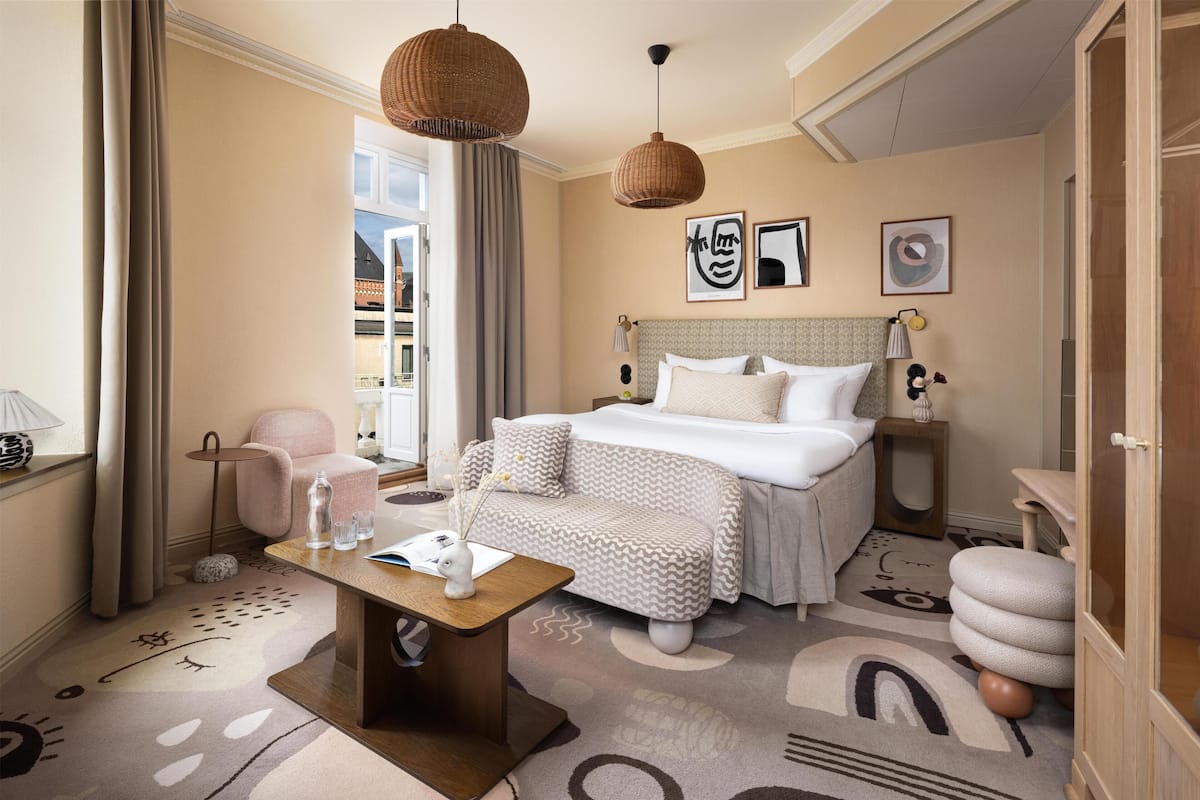 A hotel room with a bed, bedside tables, a desk, a chair, a window with curtains, and artwork on the wall.