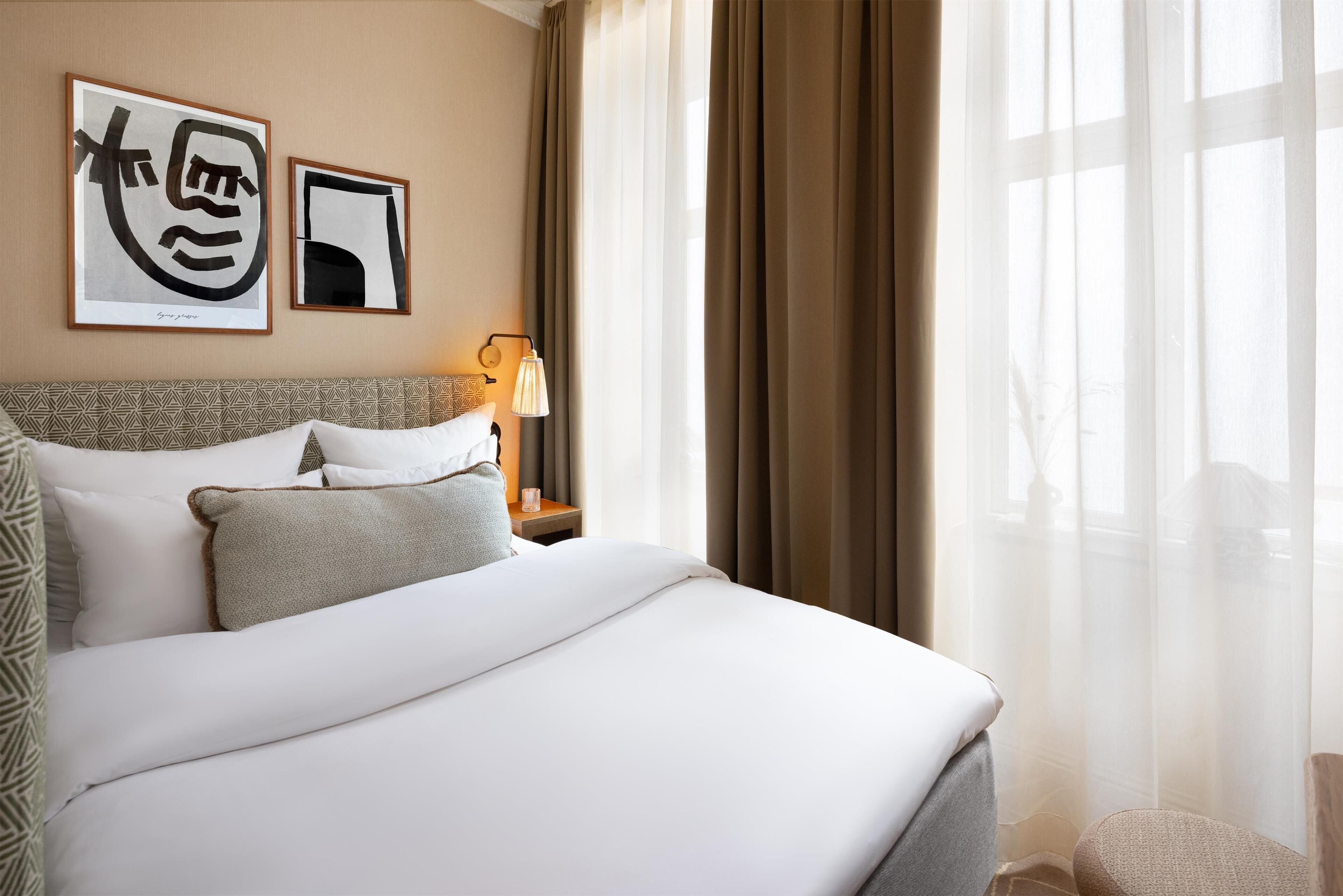 A hotel room with a neatly made bed, a bedside table with a lamp, and two framed artworks on the wall.