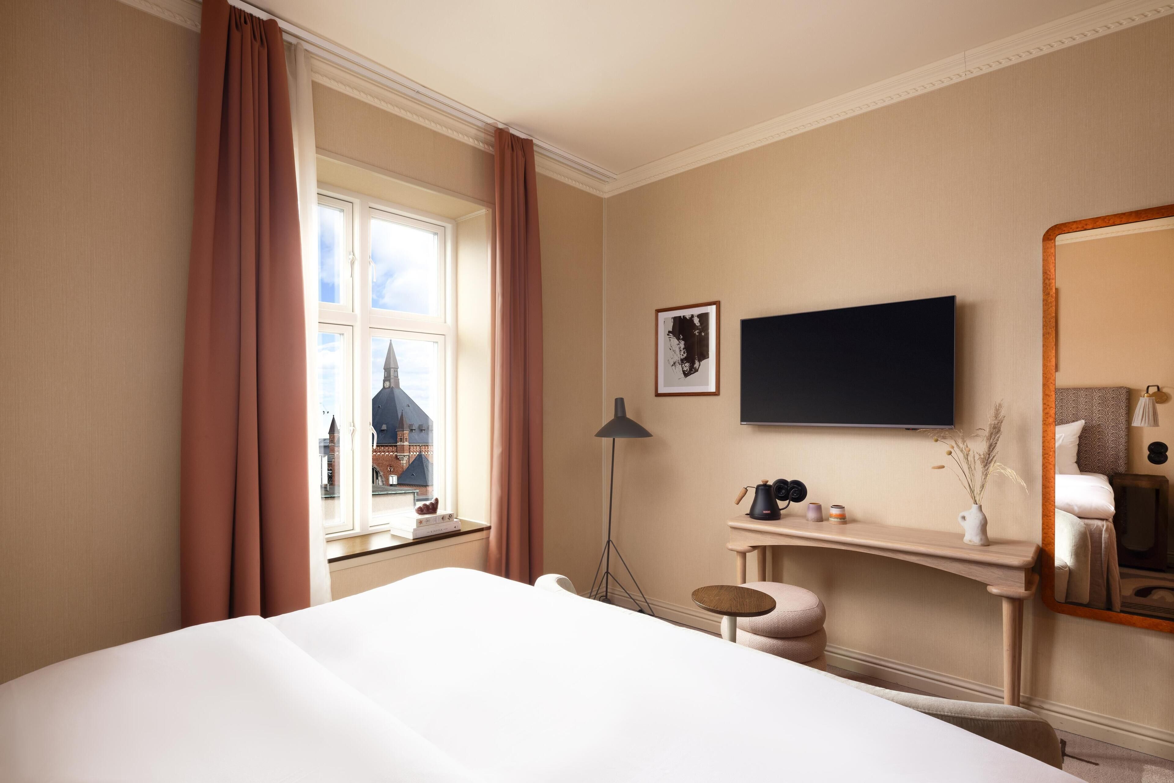 Une chambre d’hôtel avec un lit, un bureau, une chaise, une télévision et une fenêtre avec des rideaux.