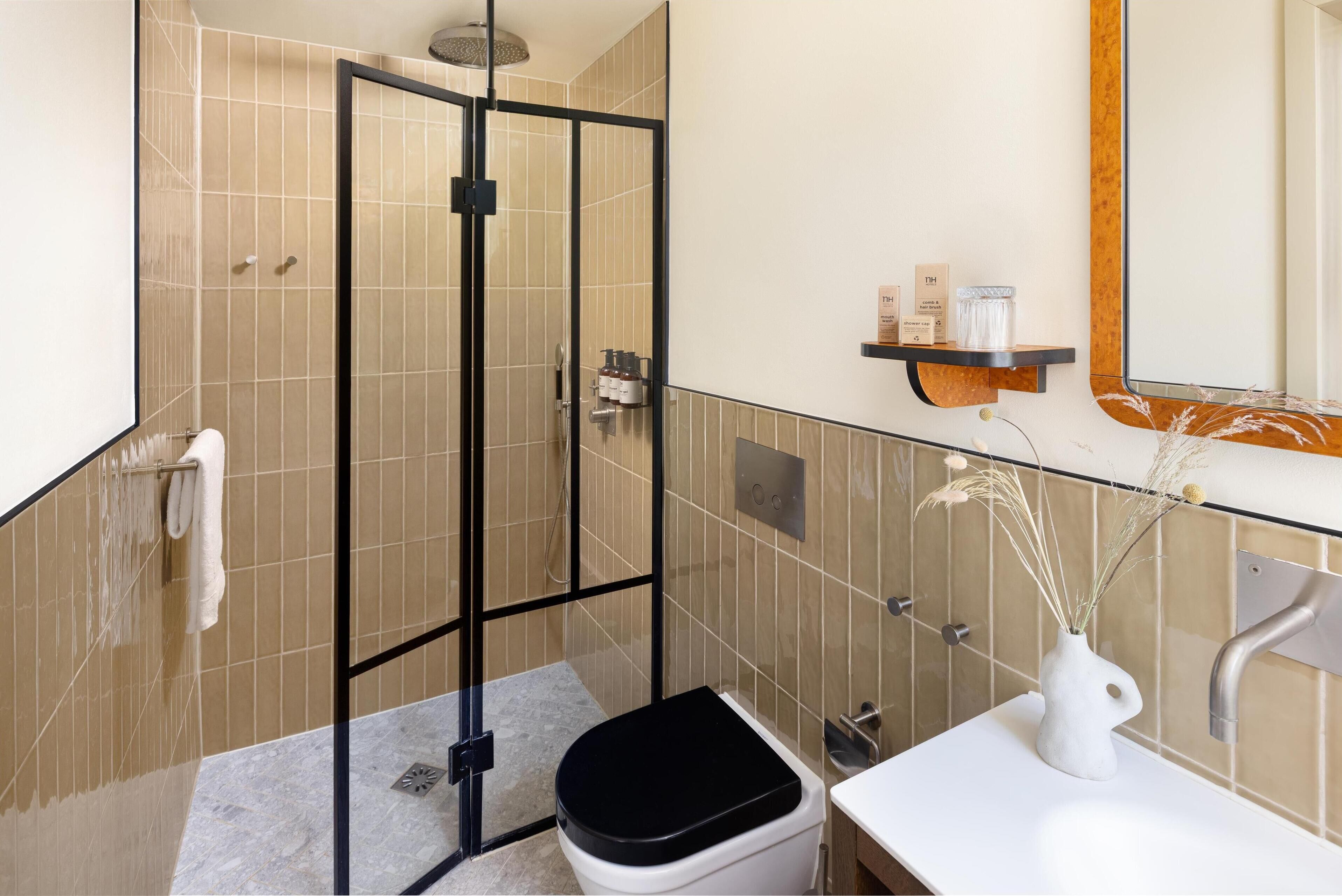 A modern bathroom with a glass-enclosed shower, a white toilet, a sink with a vase of dried plants, and a shelf with toiletries.
