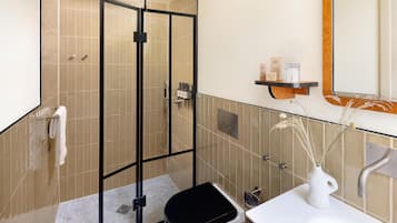 A modern bathroom with a glass-enclosed shower, a white toilet, a sink with a vase of dried plants, and a shelf with toiletries.