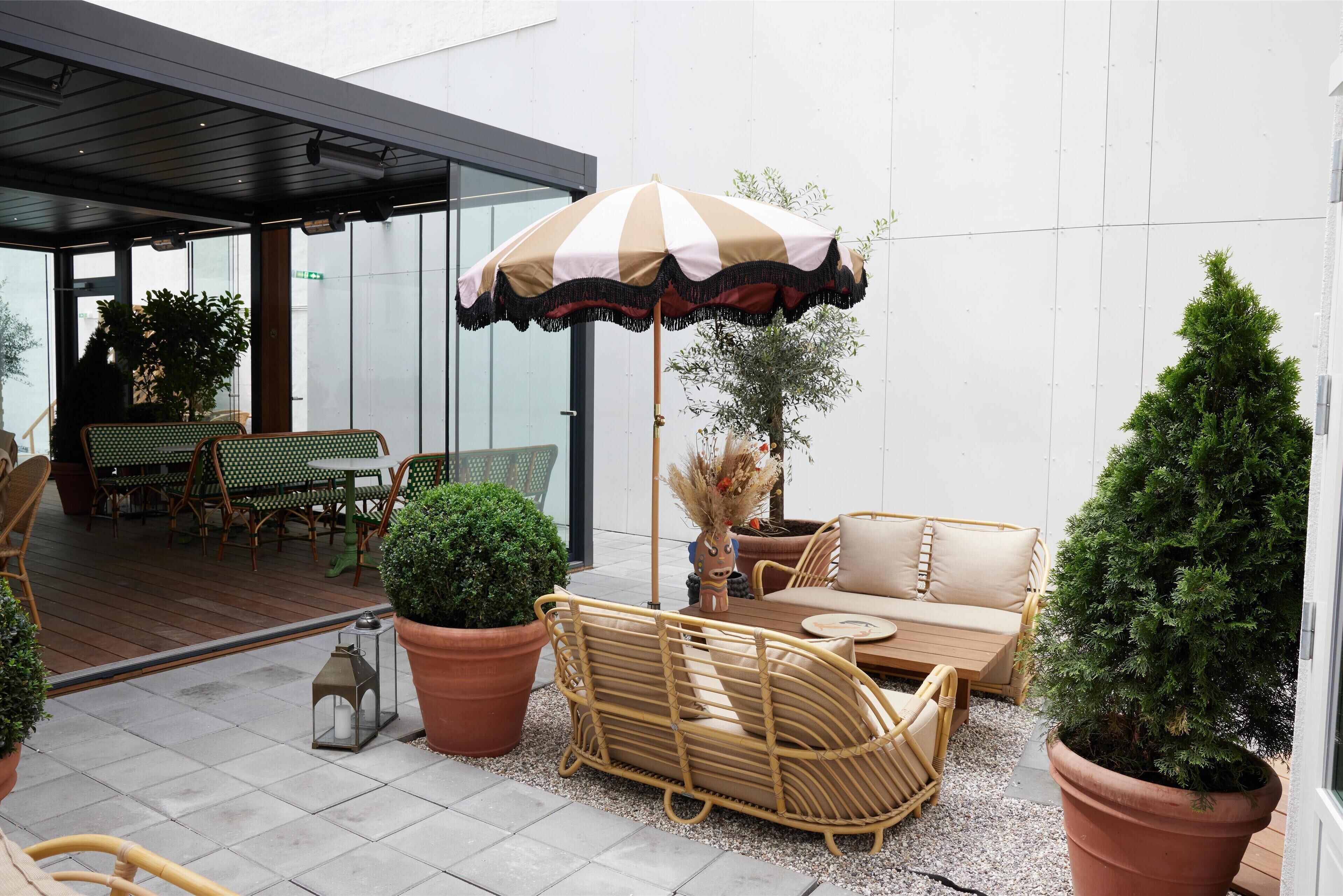 Een terras met rieten meubels, een gestreepte parasol, plantenbakken en een glazen wand op de achtergrond.