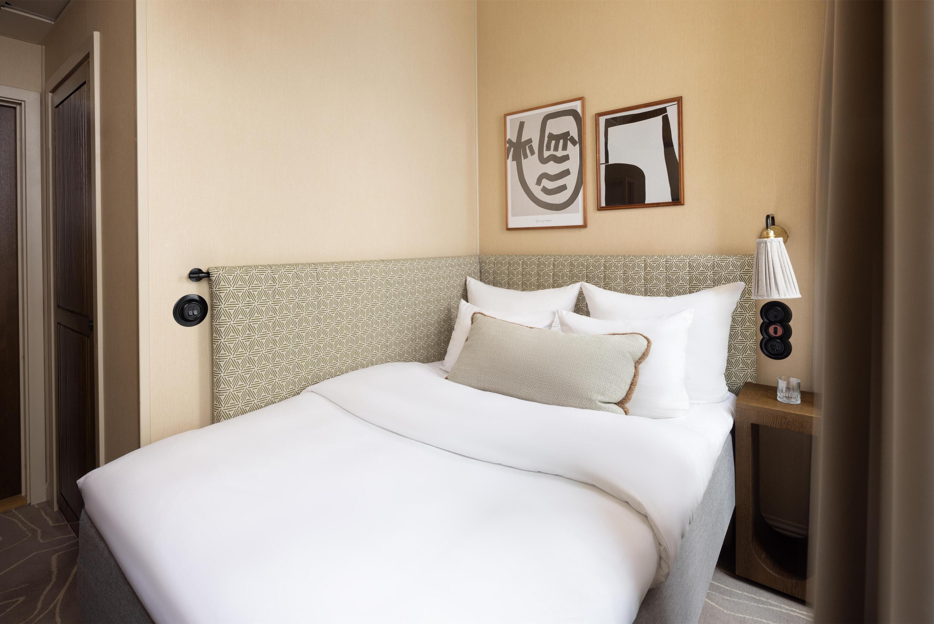 A hotel room with a large bed, a bedside table with a lamp, and two framed artworks on the wall.