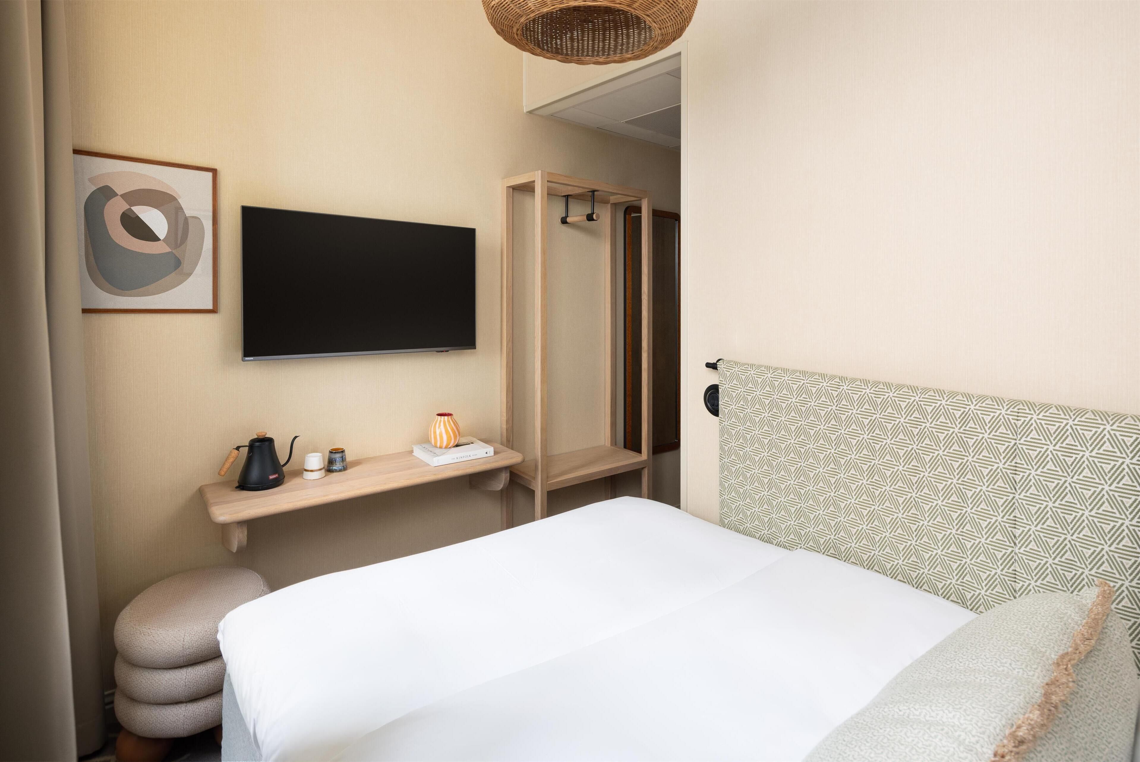 Habitación de hotel con cama, televisor montado en la pared, armario de madera y estante con tetera y un florero.
