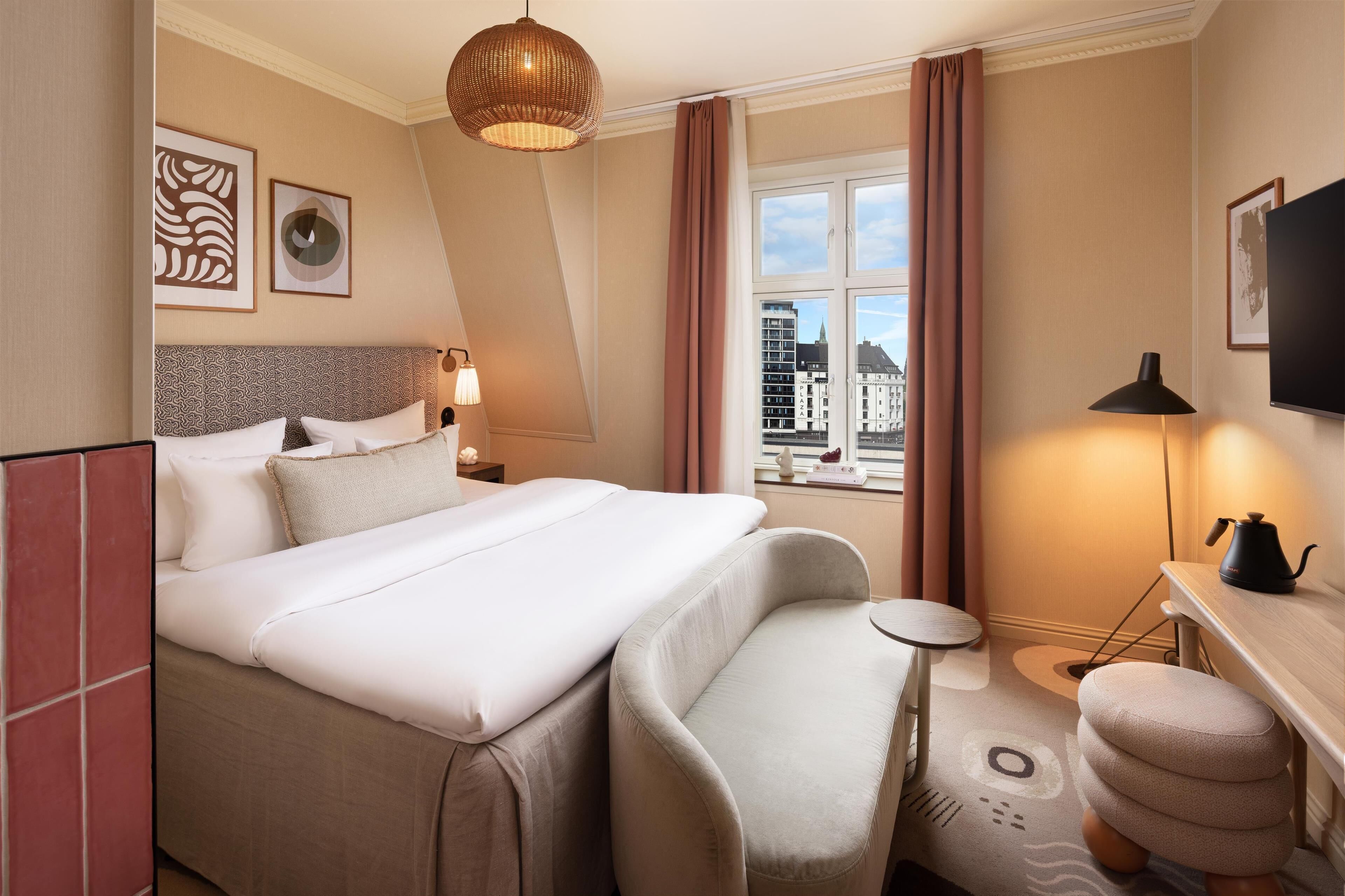 A hotel room with a large bed, a desk, a chair, a small round table, a television, and a window with a city view.