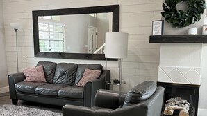 Un salon avec un canapé en cuir foncé, un fauteuil gris, un tapis à longues mèches gris, un lampadaire, un miroir et une cheminée avec des bûches.