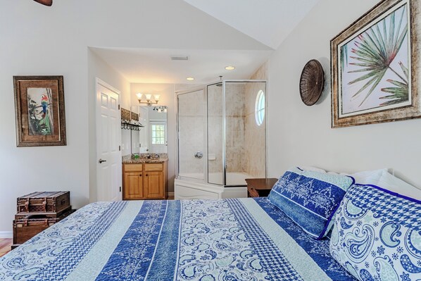 A bedroom with a large bed, a bathroom with a shower, and a hallway leading to an adjacent room.