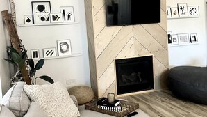 Un saló modern amb un mur amb dibuix de punt de serra, un televisor de pantalla plana i una llar de foc. Hi ha quadres a la paret, un sofà i una taula de centre amb un llibre i una espelma.