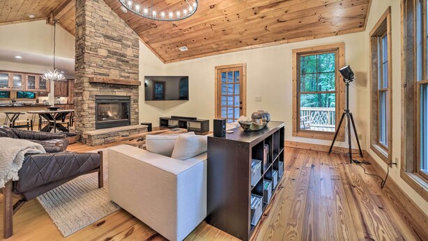 A cozy living room with a stone fireplace, wooden ceiling, and large windows offering a view of the outdoors. - Secluded Sapphire Chalet w/ Game Room + Decks (Sapphire)