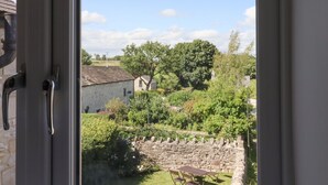 Blick durch ein Fenster auf ein Steinhaus mit Garten, Steinmauer und einem großen Baum.
