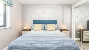 A modern bedroom with a large bed, two bedside tables with lamps, patterned wallpaper, and a sliding glass door. - 2 bedroom accommodation in Newquay (Newquay)