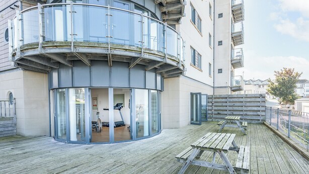 A modern building with a curved balcony, glass doors leading to an interior space with exercise equipment, and a wooden deck with a picnic table. - 2 bedroom accommodation in Newquay (Newquay)