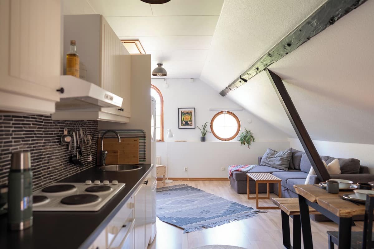 A modern kitchen with a stove, cabinets, and a brick backsplash. There is a dining area with a table and chairs, a sofa, and a rug.