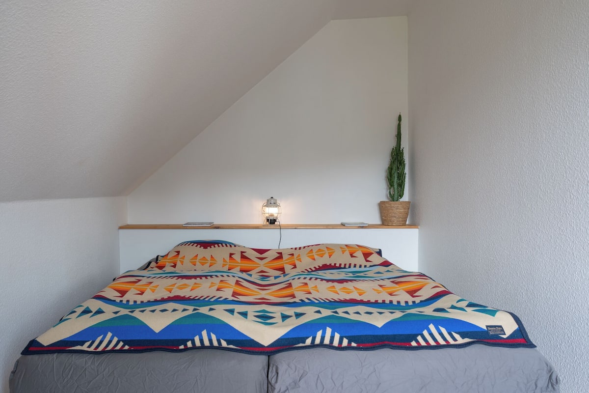 A bed with a colorful blanket, a small plant on a shelf, and a wall-mounted light fixture.