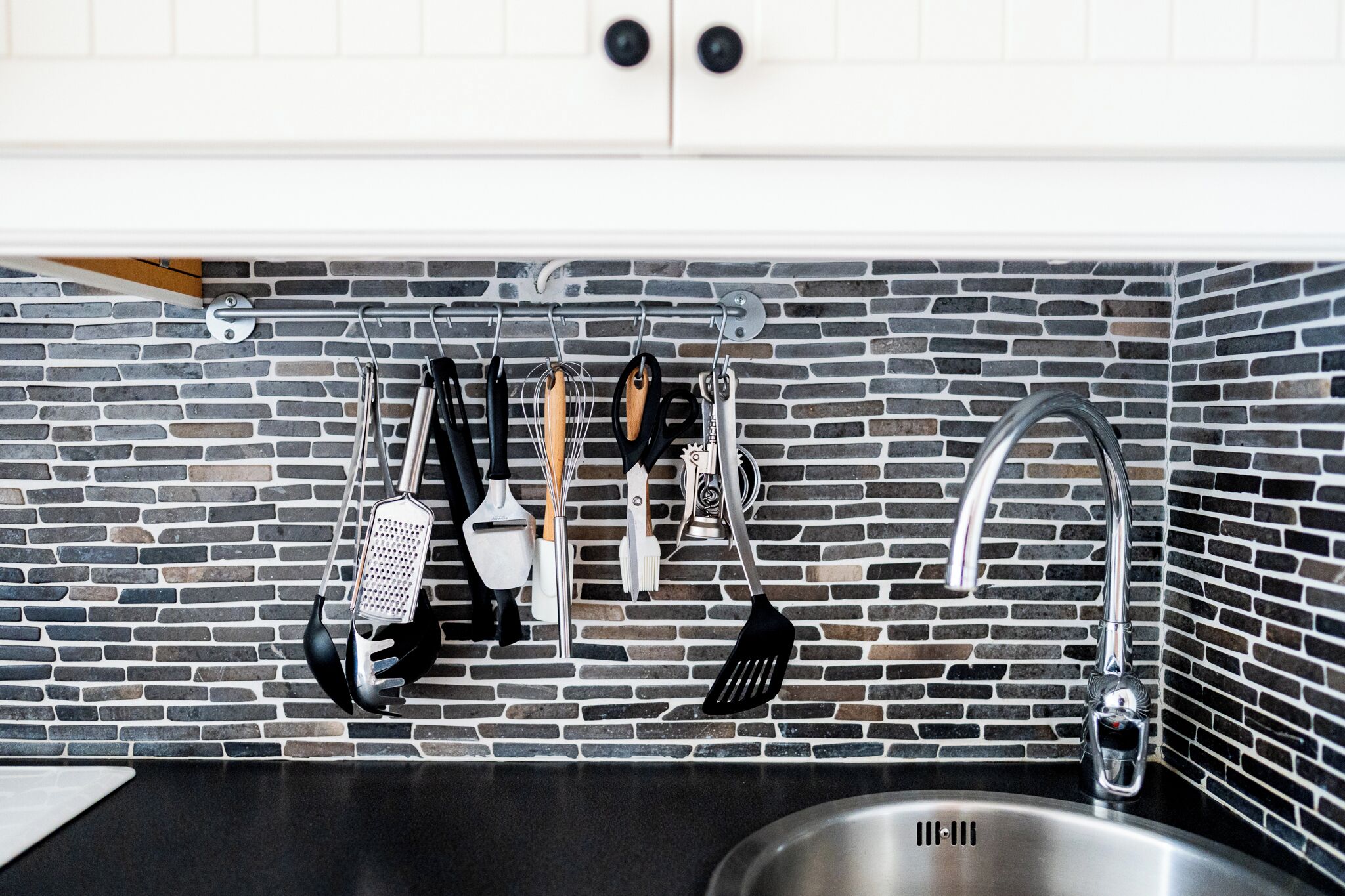 A kitchen with a black countertop, a stainless steel sink, and a hanging utensil rack against a brick backsplash.