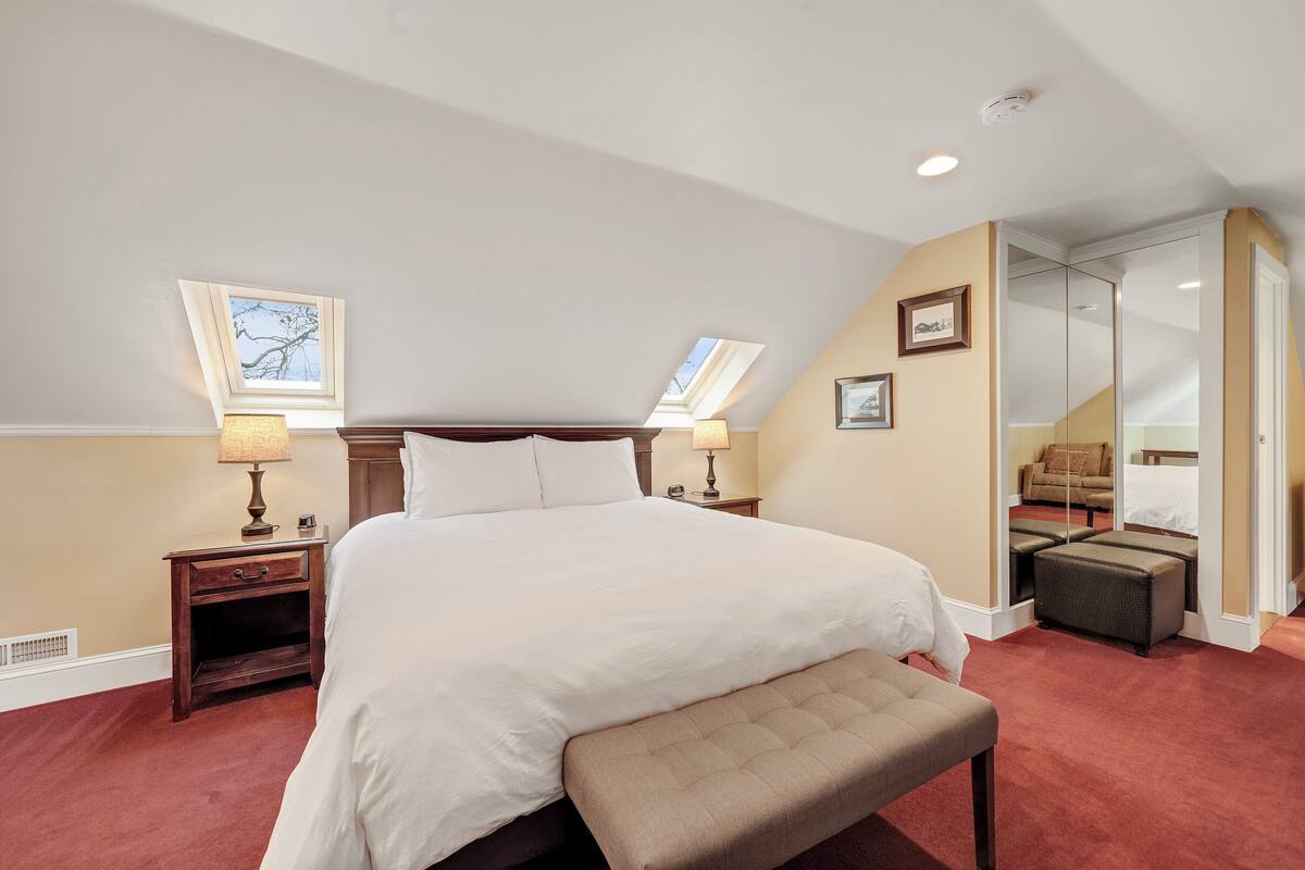 Ein Hotelzimmer mit einem großen Bett, zwei Nachttischen mit Lampen, einer Bank, einem Spiegel und Blick ins Freie durch ein Dachfenster.