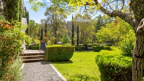 Un jardin bien entretenu, avec un chemin de gravier menant à l’entrée d’un bâtiment, le tout bordé de haies et d’arbres soigneusement taillés.