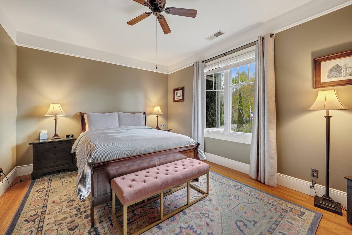 Ein Schlafzimmer mit einem Bett, Nachttischlampen, einer Bank, einem Deckenventilator und einem Fenster mit Vorhängen.