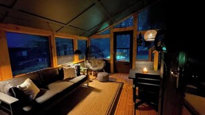 A cozy room with a sofa, a hanging chair, a table with a candle, and large windows overlooking a night scene. - Saturdays Cabin, Hot Tub, Hammock, Gas Firepit (pocono lake)