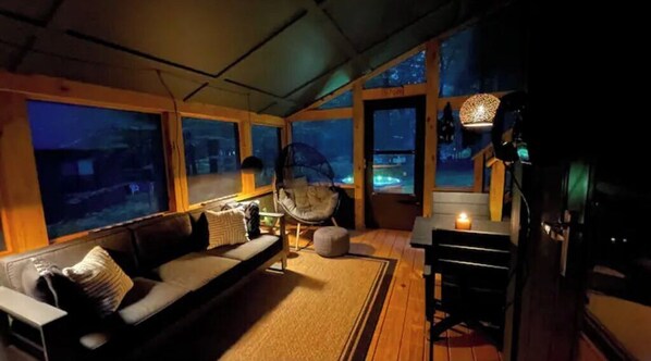 Une pièce chaleureuse avec un canapé, un fauteuil suspendu, une table avec une bougie et de grandes fenêtres donnant sur un paysage nocturne.