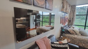 Ein modernes Wohnzimmer mit einem großen Spiegel, der einen Küchenbereich reflektiert, einer Couch mit gemusterten Kissen und Blick auf eine grüne Landschaft durch große Fenster.