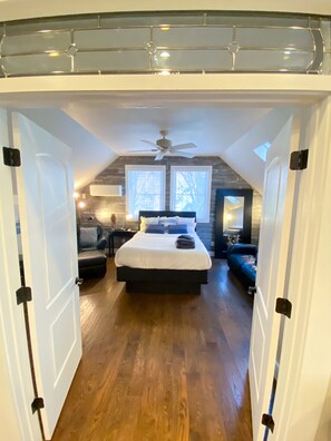 Une chambre avec un grand lit, un ventilateur de plafond et une vue sur l’extérieur grâce à deux fenêtres.