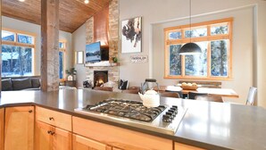 Una cocina moderna con una isla grande, armarios de madera, chimenea de piedra y amplios ventanales con vistas al exterior.