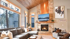 Una sala moderna con chimenea de piedra, un televisor de pantalla plana grande montado encima, y un área de descanso acogedora con un sofá y sillones.