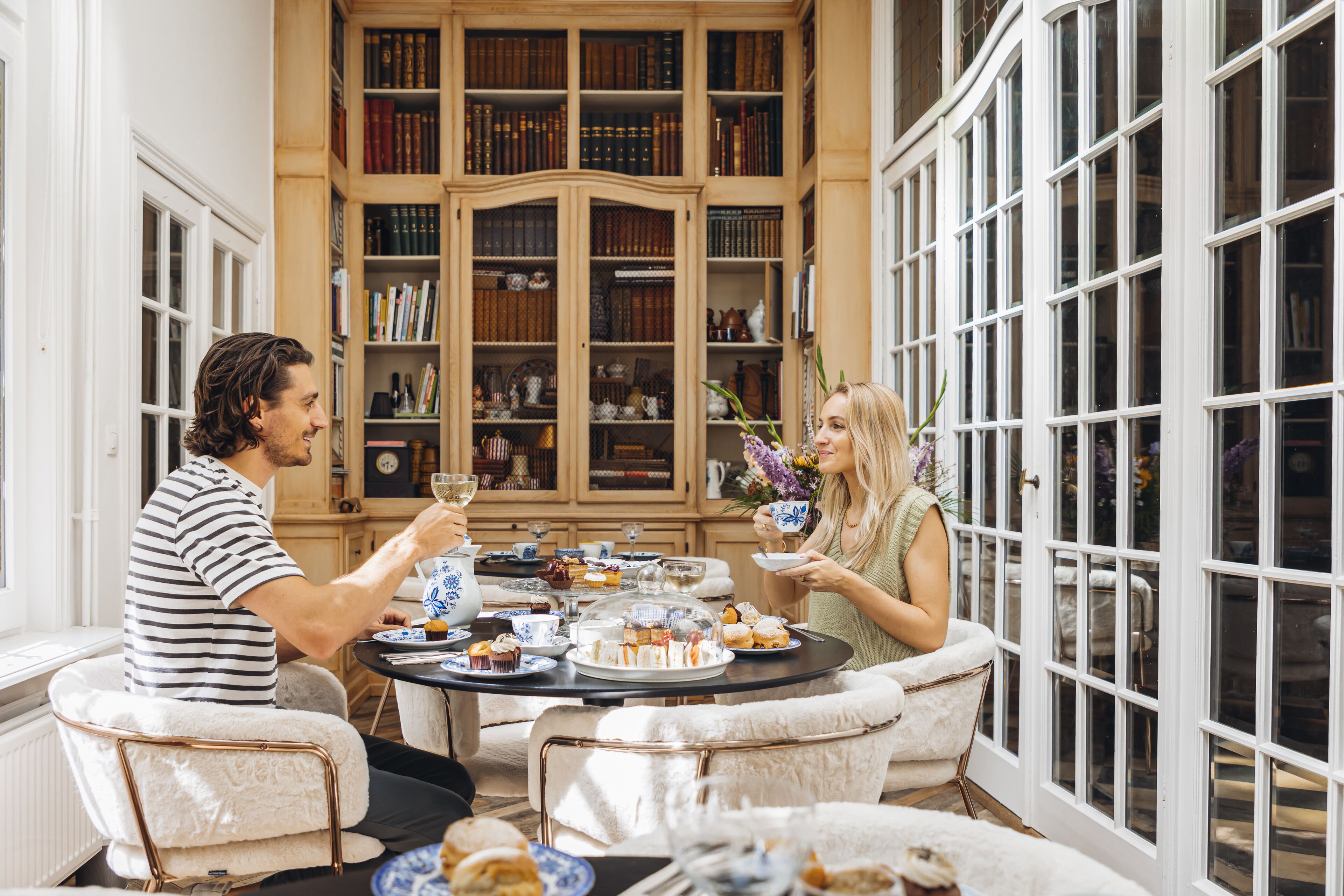 Een paar zit aan een tafel met verschillende gebakjes en drankjes, omringd door boekenplanken en grote ramen.