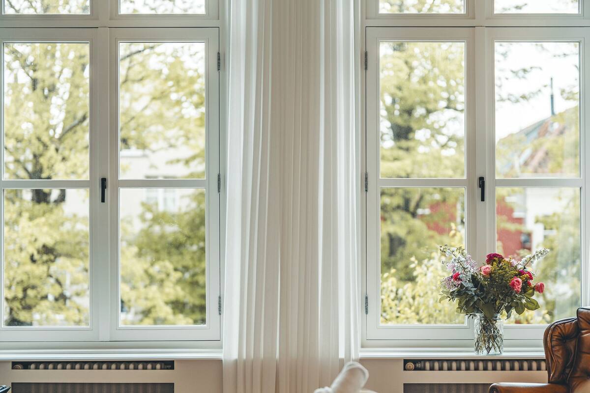 Ett sovrum med en säng, ett fönster med gardiner, ett element och en vas med blommor.