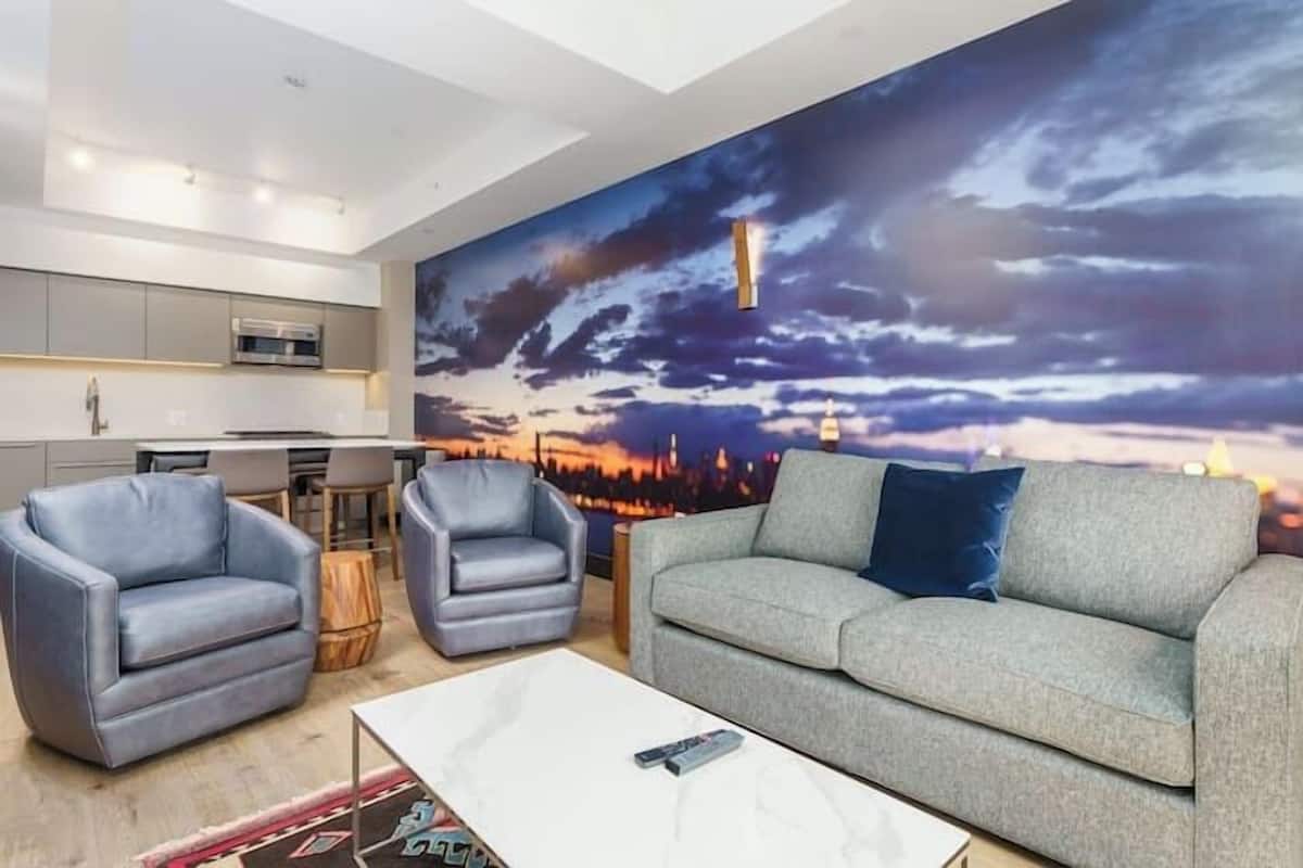 Uma sala de estar moderna com um sofá cinza, duas poltronas e uma mesa de centro. Um mural com vista para a cidade está na parede atrás do sofá.