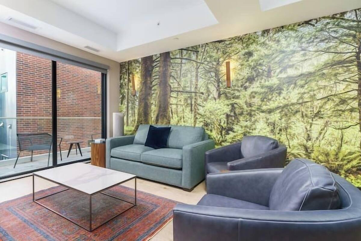 Uma sala de estar moderna com uma janela grande, um sofá verde, uma poltrona de couro preta e um tapete estampado.
