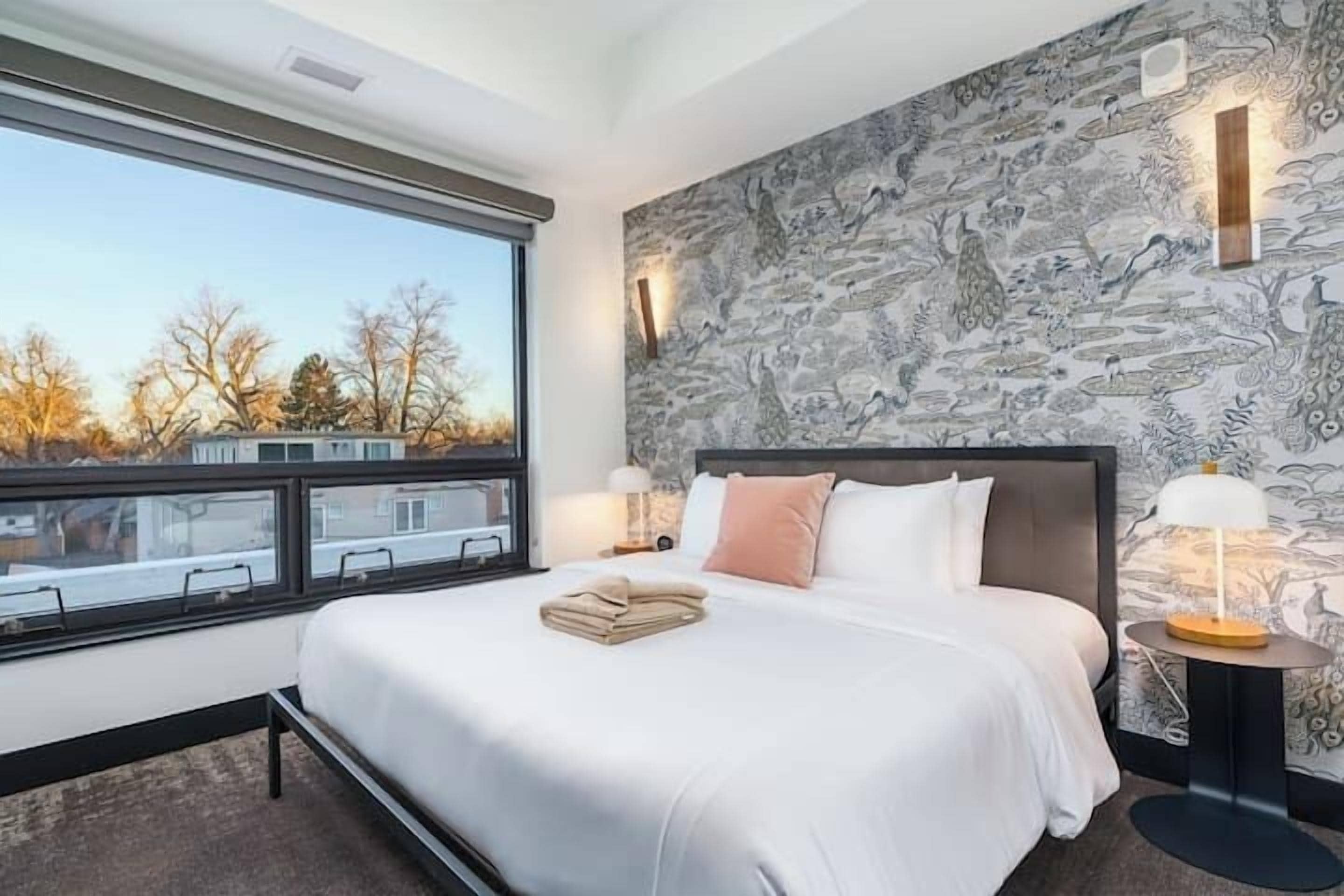 A modern hotel room with a large bed, a view of the outside, and a patterned wallpaper.
