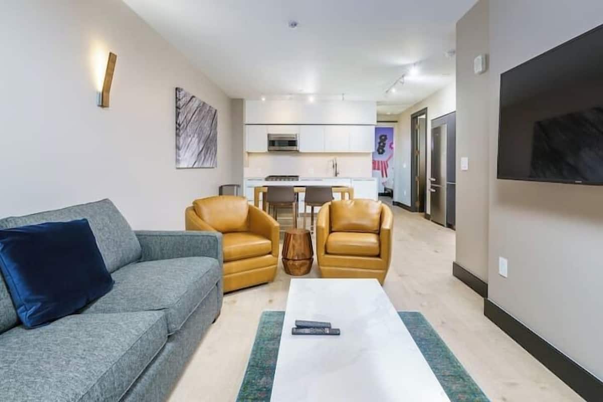 Uma sala de estar moderna com um sofá cinza, duas poltronas amarelas, uma mesa de centro branca e uma TV fixada na parede.
