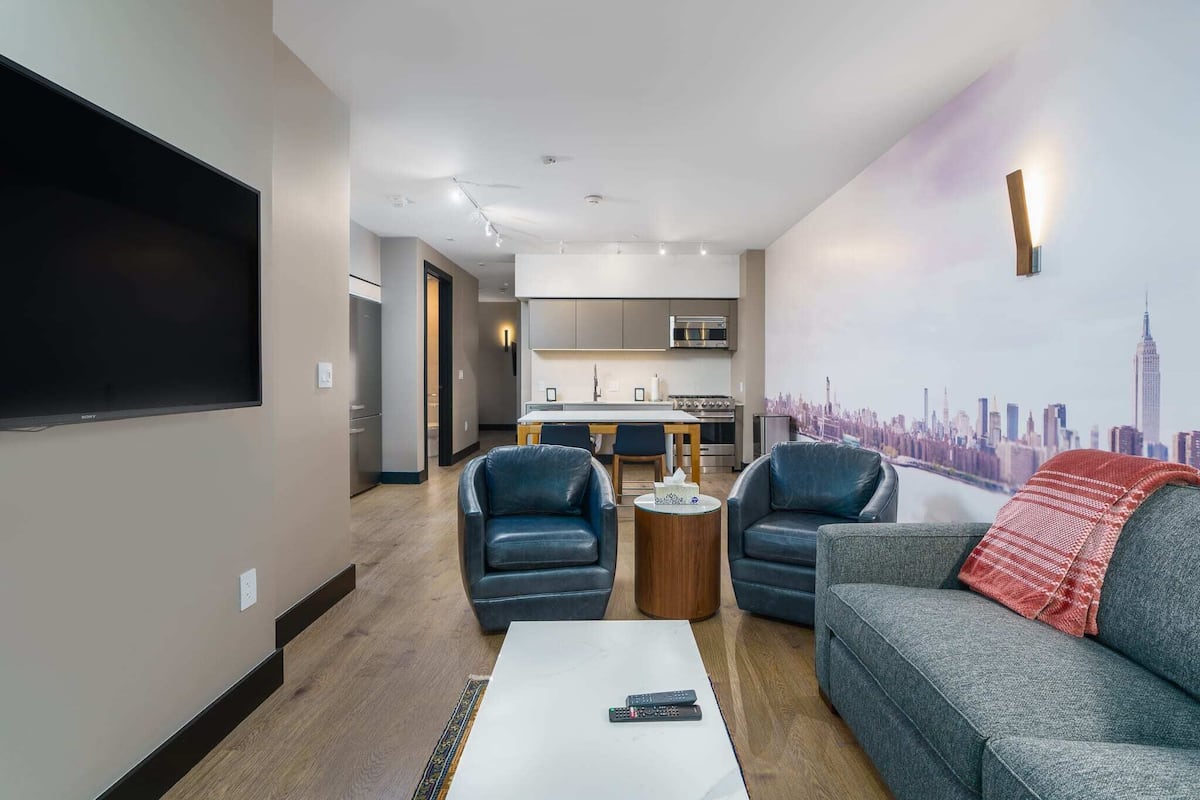 Uma sala de estar moderna com uma TV de tela plana, duas poltronas de couro, um sofá cinza e uma mesa de centro. Uma cozinha é visível ao fundo, com um mural de paisagem urbana na parede.