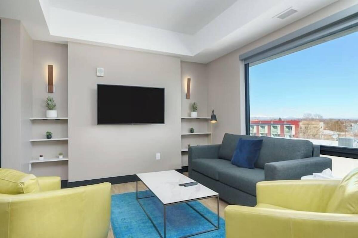 Uma sala de estar moderna com uma TV de tela plana na parede, um sofá cinza com almofadas azuis, uma poltrona amarela e um tapete azul.