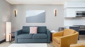 A modern living room with a grey sofa, a tan armchair, and a white coffee table. A kitchen with stainless steel appliances is visible in the background.