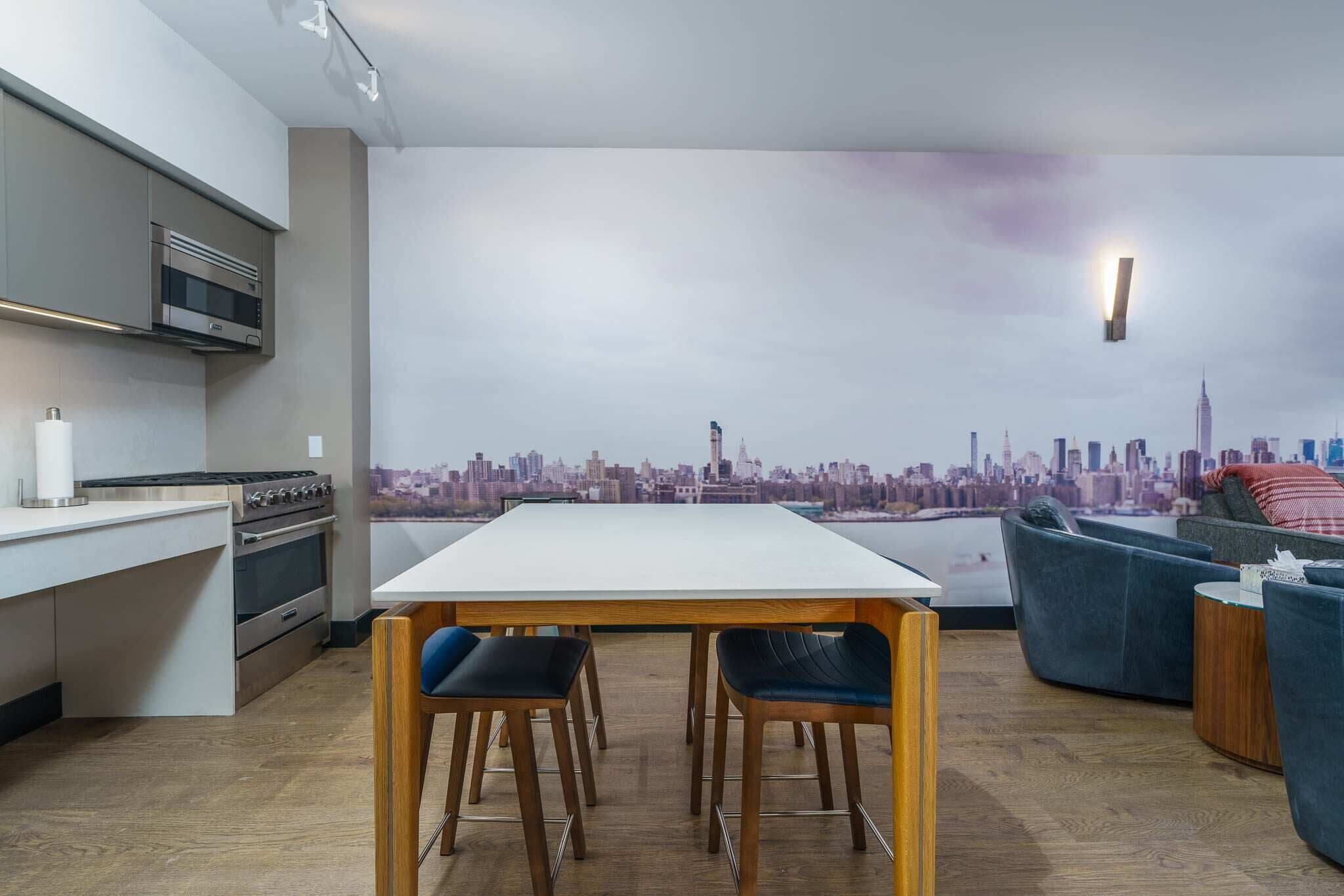 Une cuisine moderne ornée d’une fresque représentant un paysage urbain, un coin repas avec une table blanche et des chaises bleues, et un canapé bleu avec un coussin rouge.