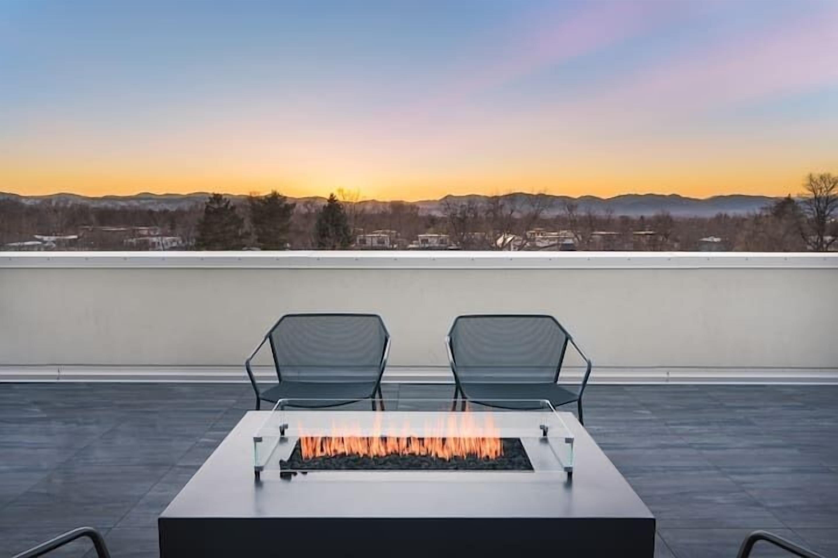 Une terrasse sur le toit, dotée d’un foyer et de deux chaises, offrant une vue sur le paysage urbain au coucher du soleil.