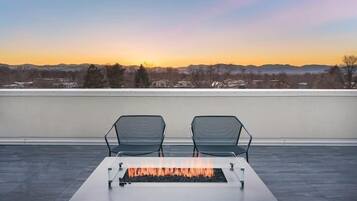 Une terrasse sur le toit, dotée d’un foyer et de deux chaises, offrant une vue sur le paysage urbain au coucher du soleil.