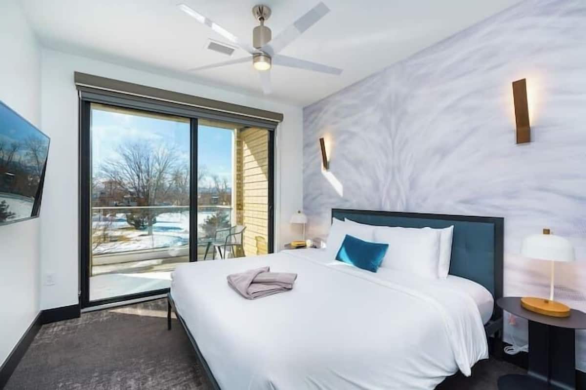 Quarto de hotel moderno com uma cama grande, um ventilador de teto, vista para o exterior e uma parede de mármore.