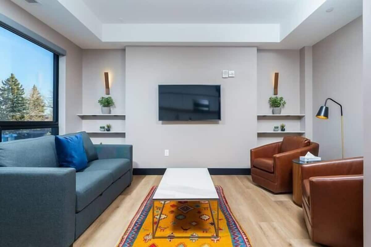 Uma sala de estar moderna com um sofá cinzento, uma poltrona de couro castanho, uma mesa de centro branca e um tapete colorido. Há uma televisão de tela plana na parede e duas pequenas prateleiras com plantas.