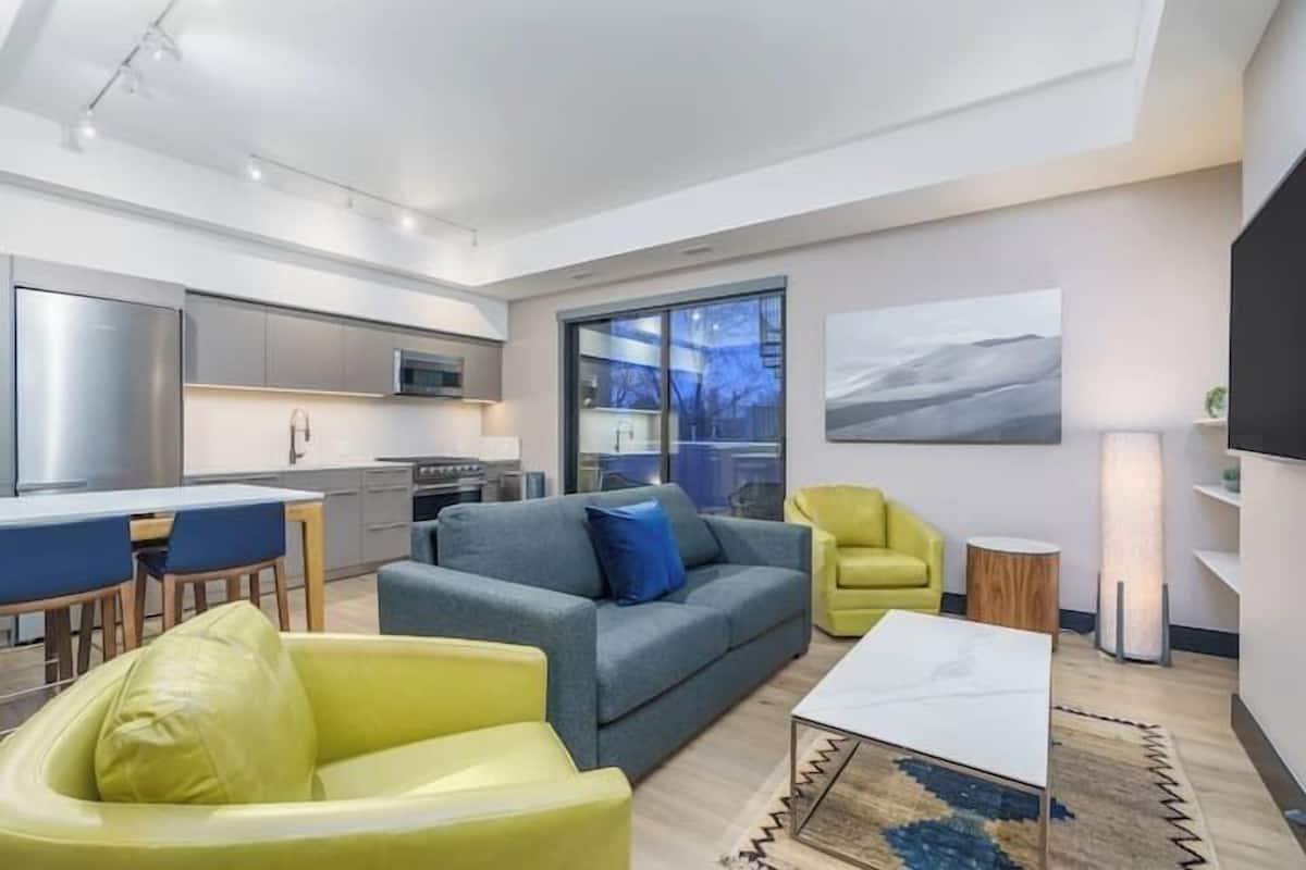 Uma sala de estar moderna com sofá, poltrona e mesa de centro. Há uma cozinha com geladeira, micro-ondas e armários. Uma grande janela oferece vista para o exterior.