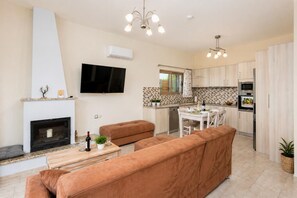 A modern living room with a fireplace, sofa, TV, and dining area with a kitchen in the background. - Stefania Villa Dio - One Bedroom Villa, Sleeps 2 (Roumeli)