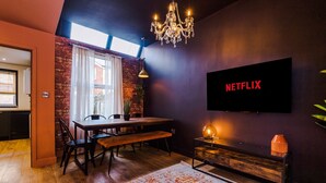 A modern living room with a dining area, a chandelier, a TV mounted on the wall, and a large window with curtains. - 4 Bed Urban Home in Manchester (Manchester)