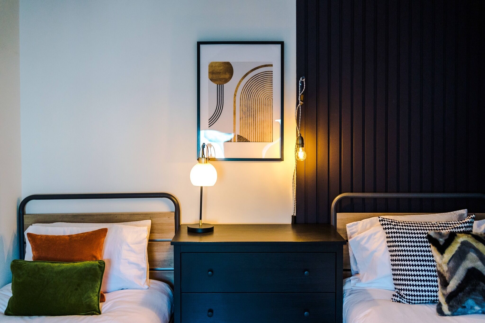 Habitación de hotel con dos camas, una mesita de noche con lámpara, un cuadro enmarcado en la pared y cabecero de madera.
